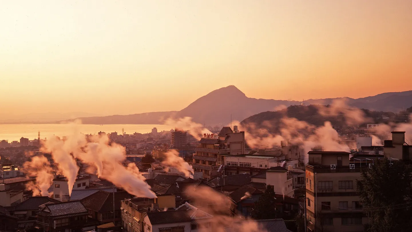 Steam rising from hot-spring vents between the houses of Beppu, in Ōita, at dusk, with the bay in the distance