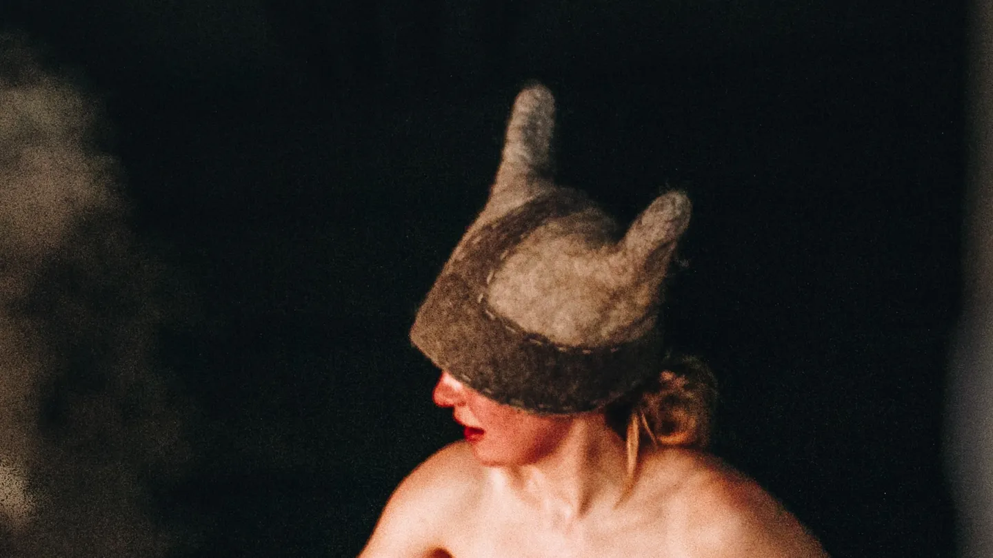 A bather in a horned felt sauna hat in an Estonian smoke sauna, with steam rising from the stones alongside
