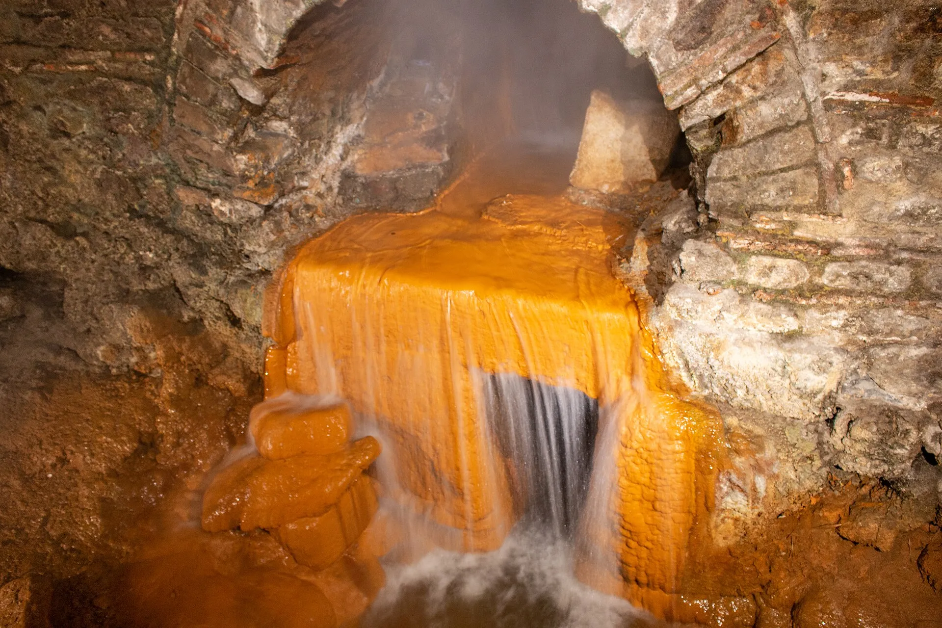 Hot water from the Sacred Spring at Bath running over stones stained orange by iron, under a stone arch in the Roman Baths