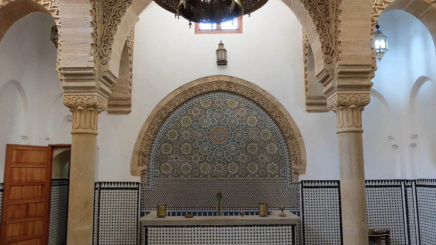 Reception hall of the Hammam Saffarin in Fez, with carved stucco arches, a mosaic wall fountain and a checkered floor