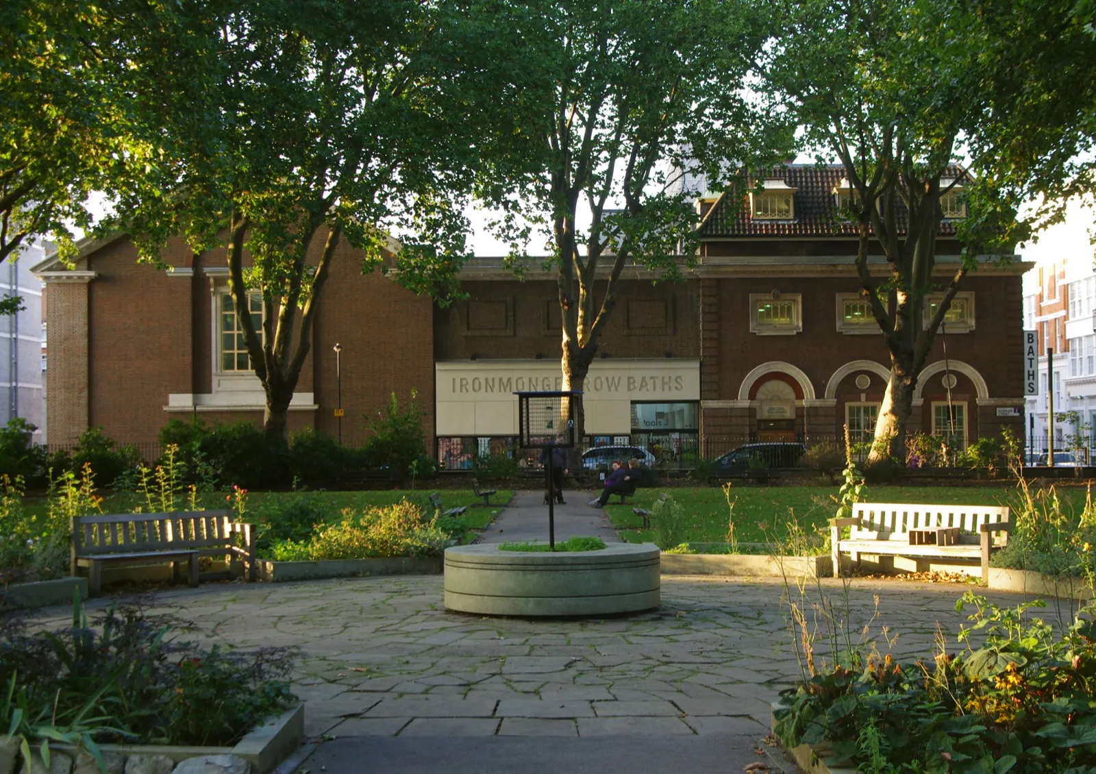 Brick facade of Ironmonger Row Baths seen from the garden behind St Luke's church, with trees and benches