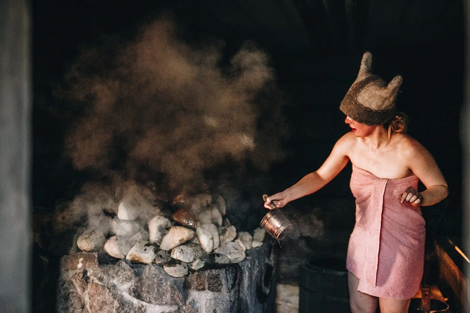 Woman in a towel and felt hat throwing water on the stones of a smoke sauna in Estonia, with steam rising