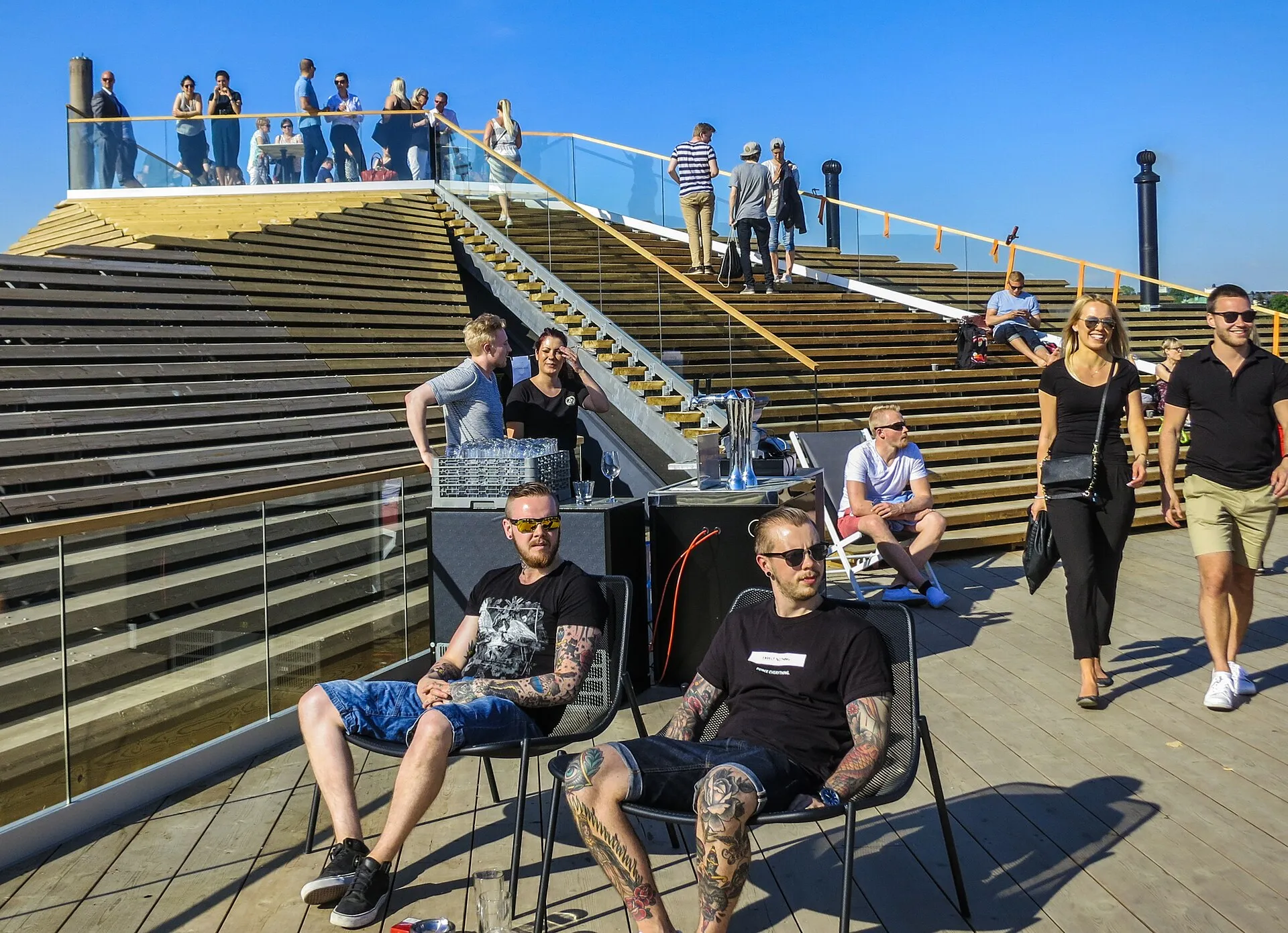 Timber terrace at Löyly in Helsinki, with people seated in chairs and standing on the steps of the tiered roof under a blue sky