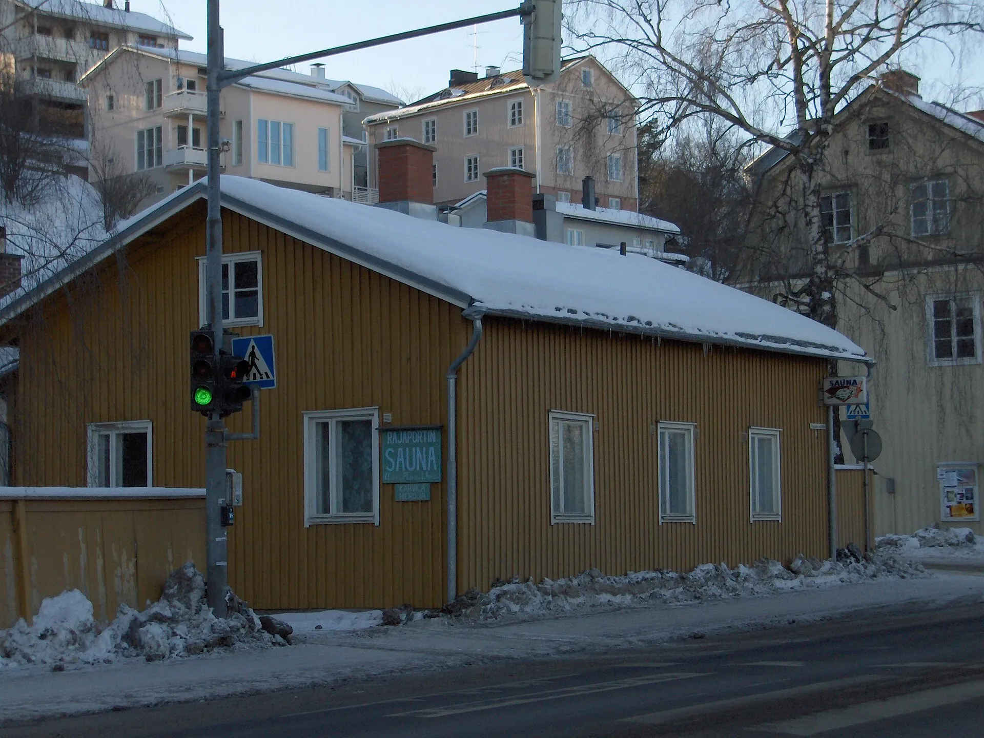 Low yellow timber building on the corner of a snowy street in Pispala, Tampere, with the Rajaportti sauna sign on its facade