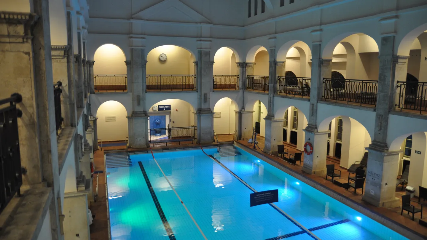 Empty indoor swimming pool at Rudas Baths in Budapest, with illuminated blue water and two levels of arched galleries