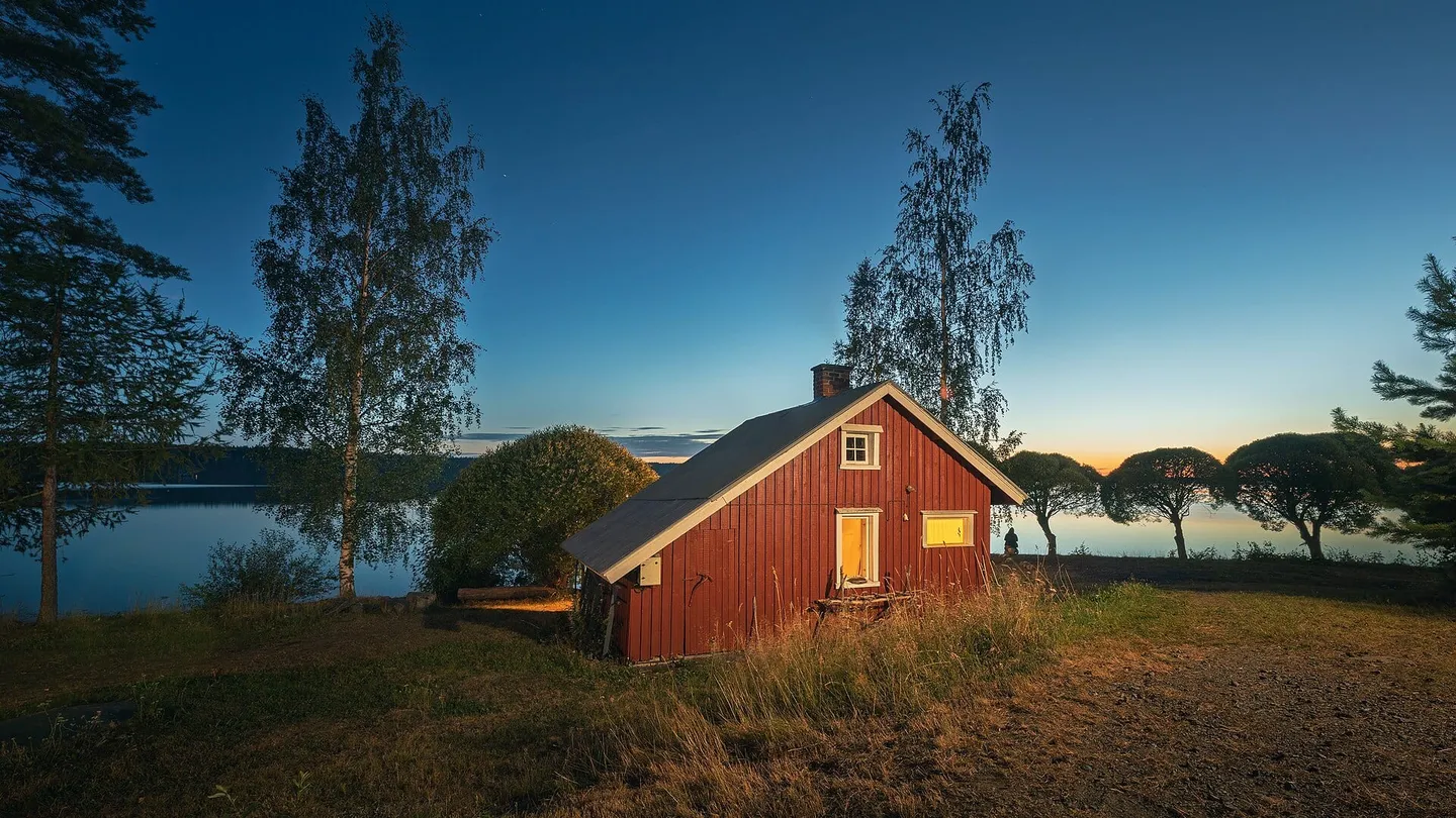 Red timber sauna cabin on the shore of Lake Ätäskö in Kitee, Finland, with its light on at dusk