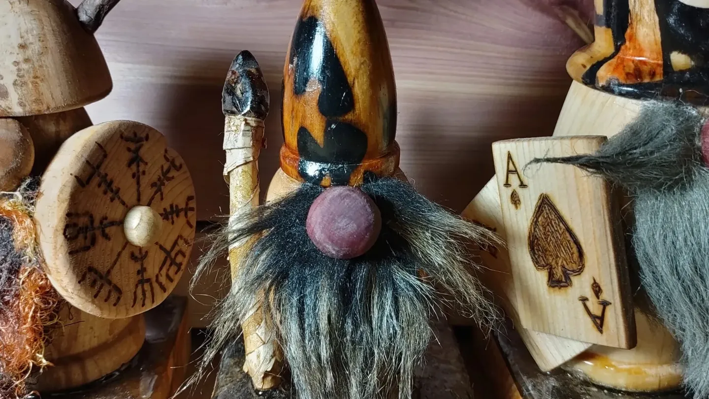 Three carved wooden saunatonttu with fur beards on a wooden shelf; the one in the middle, with a pointed hat and a round nose, holds a staff