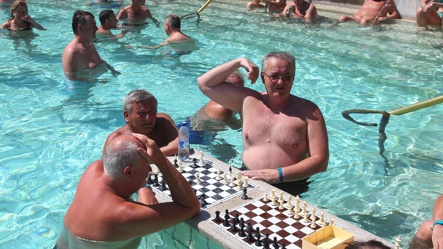 Men playing chess on a board resting on the edge of the outdoor thermal pool at the Széchenyi Baths in Budapest, surrounded by dozens of bathers
